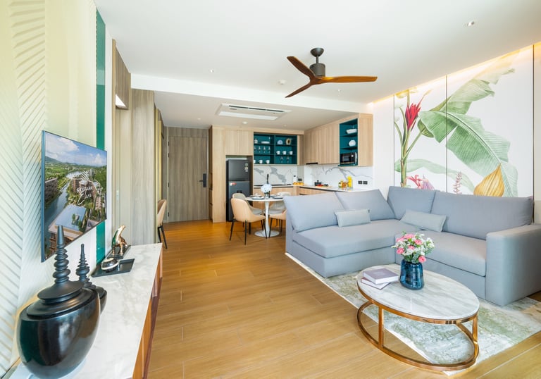 Modern open-plan living room with blue sectional sofa, wooden floors, kitchen area, and decorative plants near windows with tropical artwork