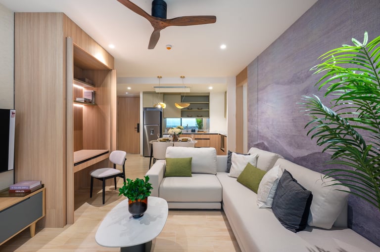 Modern living room with beige sectional sofa, wooden desk nook, purple accent wall, potted plant, and open kitchen visible in background