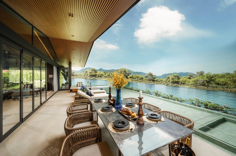 Modern riverside deck with dining table, wicker chairs, and glass railings overlooking a river and lush mountains