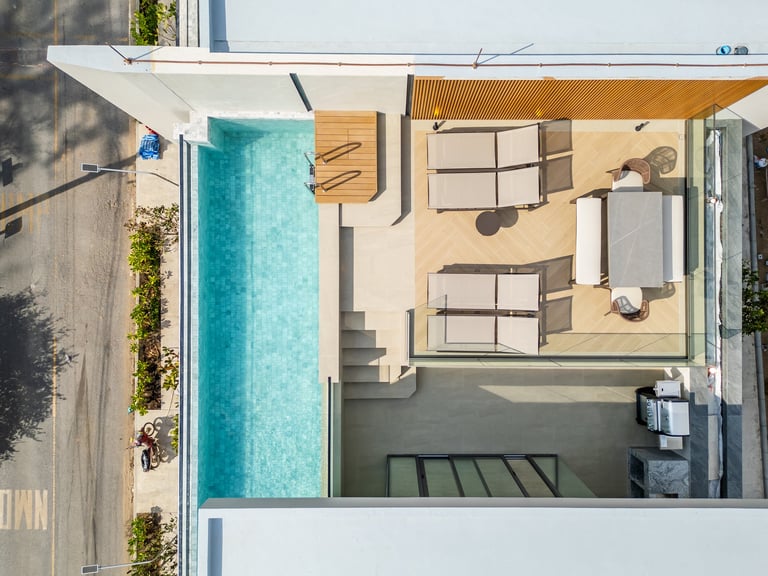 Overhead view of a modern house with a turquoise swimming pool, wooden deck, sandy patio, and furnished living space